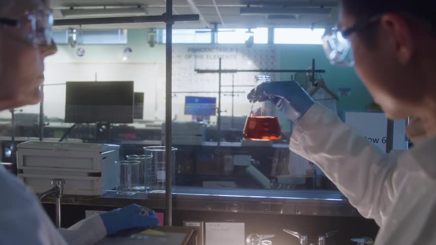 The video shows two scientists in a lab wearing lab coats and safety glasses looking at a chemical reactionthe scientist on the right wearing blue gloves is holding up an erlenmeyer flask containing a bright orangered liquid for ex