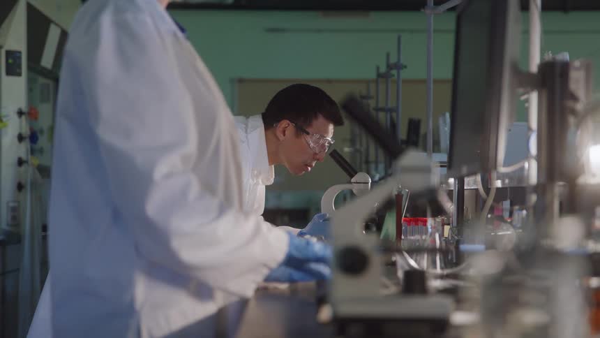 Young male scientist looking at a microscope on a lab next to another scientist indoors during day time