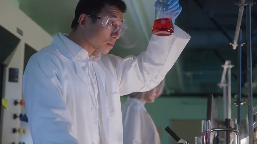 A young man in a laboratory wearing goggles and a lab coat stands beside a young woman similarly dressed he carefully inspects a red liquid contained in a beaker