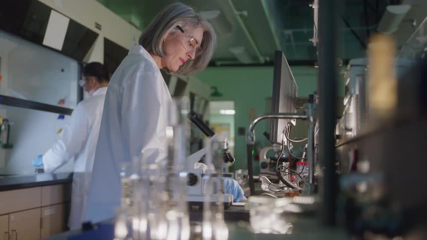 A woman scientist wearing safety glasses and blue gloves leans toward a computer beside a microscope with a focused expression while another lab worker in the background adjusts their goggles amid glassware and fume hoods