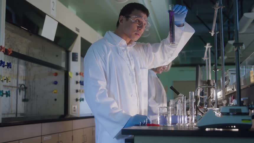 Scientist conducts an experiment with test tubes while a colleague watches in the background