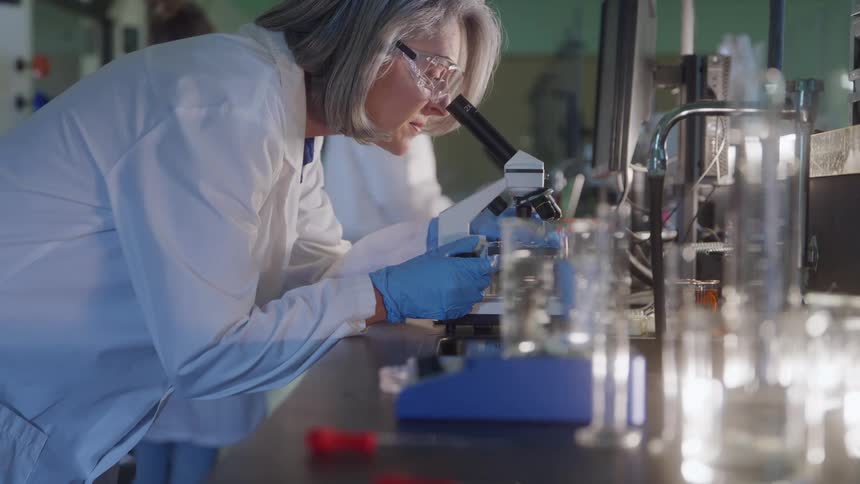 A young woman with blonde hair wearing glasses and a lab coat examines a specimen through a microscope in a laboratory