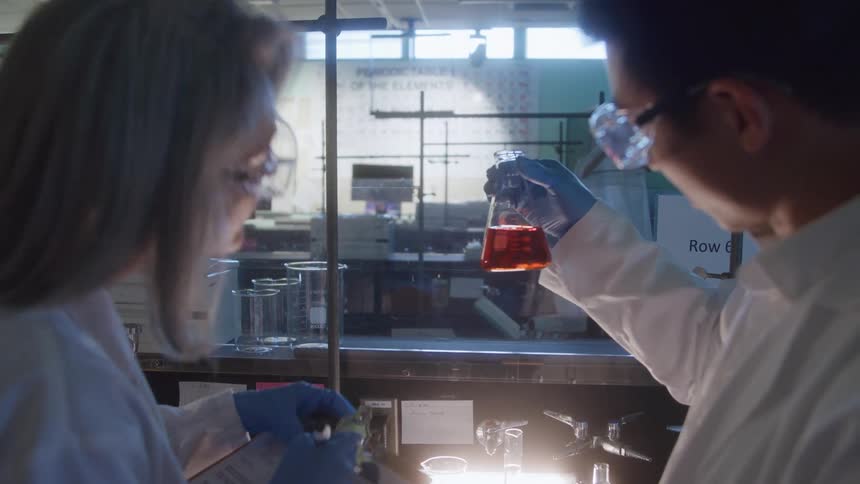 Young male scientist holding a glass flask with a red liquid while a female scientist writes down notes indoors during day time