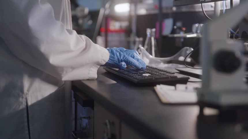 The video is a closeup of a person wearing a lab coat and blue gloves typing on a black computer keyboardthe work is being done on a countertop in a laboratory with a microscope visible on the right side of the frame