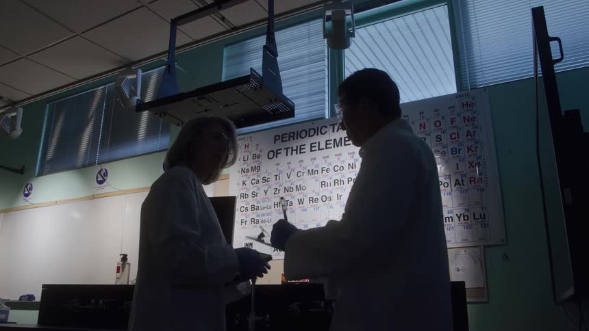 A male scientist and a young woman both wearing lab coats and goggles engage in a discussion inside the laboratory