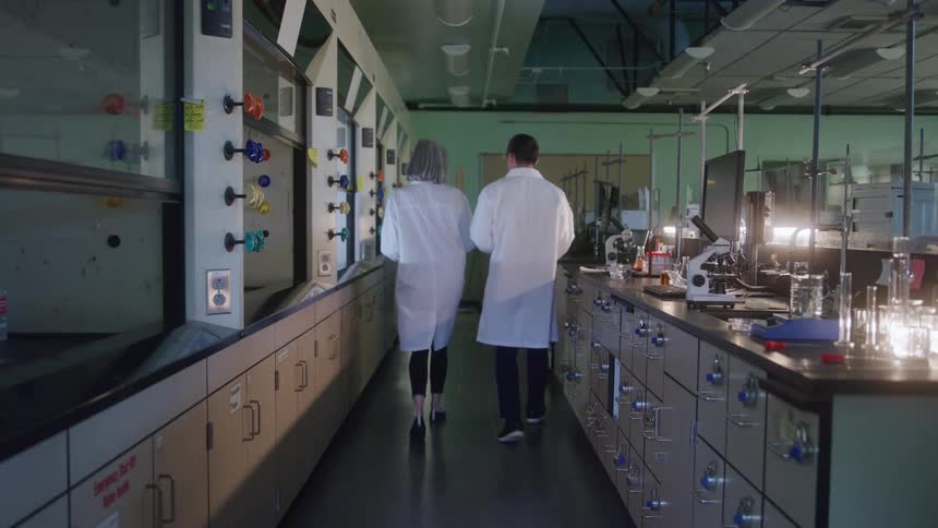 Man and woman walking through the central aisle of a modern laboratory equipped with fume hoods computers and scientific instruments