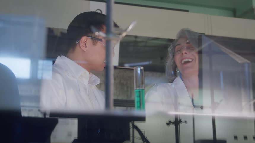 Two scientists in a lab happily discuss the results of an experiment while observing a test tube filled with a green liquid