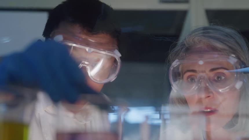 The video is a closeup of two scientistsa man and a womanboth wearing safety goggles and lab coatsthey are leaning in over glassware on a lab bench observing a reaction or sample with focused engaged expressions