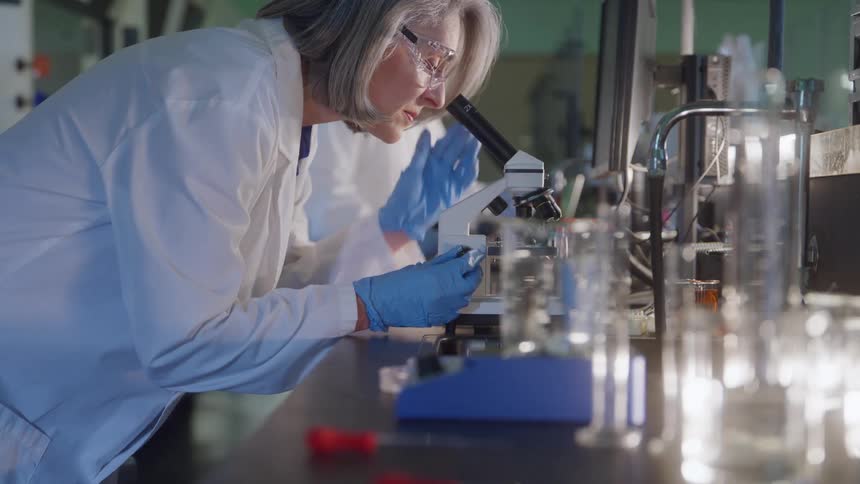A female scientist in a white lab coat and safety glasses looks through a microscope in a laboratory
