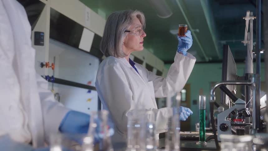 The video shows a female scientist in a lab coat and safety glasses holding up a test tube containing an amber or brown liquid for examinationshe is working at a lab bench with a microscope nearby and another person in a lab coat is partia