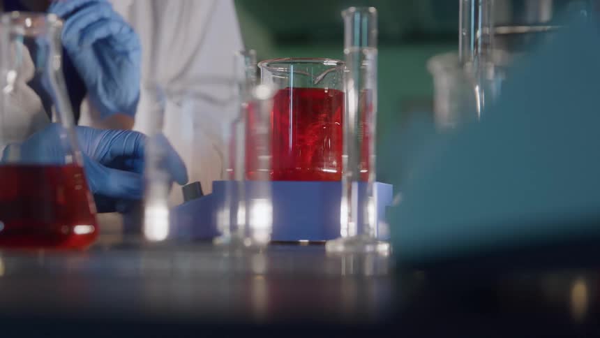 A lab scientist wearing a white lab coat and blue gloves carefully prepares a substance in a beaker