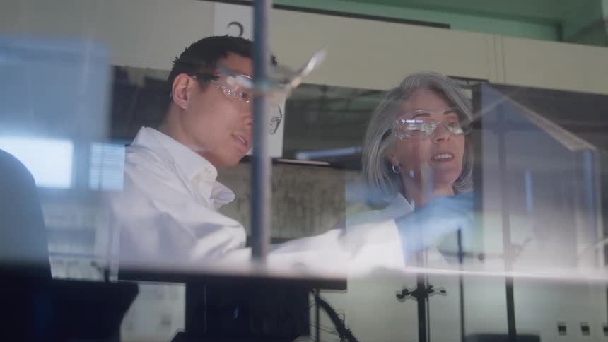 The video shows two scientists a man and an older woman standing sidebyside in a laboratory both wearing safety goggles and lab coatsthe woman appears to be speaking or explaining something while gesturing with a bluegloved hand poss