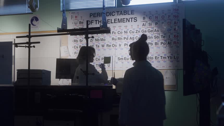The video shows a group of scientists in a lab mostly in silhouette standing in front of a large periodic table of the elementstwo people in the foreground wearing lab coats appear to be conversing and possibly reviewing notes near a la