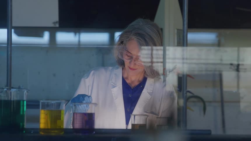 A young woman wearing a lab coat and goggles carefully takes samples from a beaker in a laboratory setting