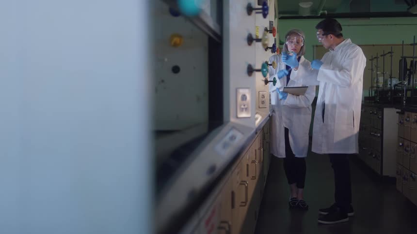 A man and a woman both wearing white lab coats safety glasses and blue gloves stand next to a large piece of laboratory equipment while inspecting a small glass vial