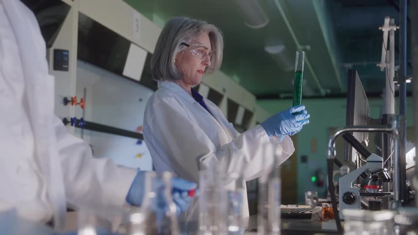 Woman analyzing green liquid in a graduated cylinder in a modern laboratory with scientific equipment and a computer in the background