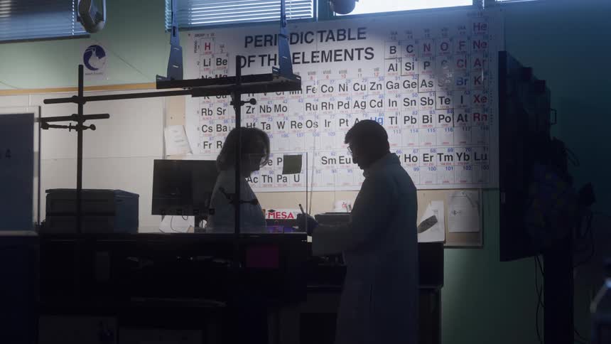 The video shows the silhouettes of two people in lab coats discussing something in a science laba large periodic table of the elements is brightly lit on the wall in the background contrasting with the dark figures in the foreground