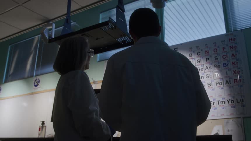 A woman and a man in white lab coats stand silhouetted in a laboratory facing a wallmounted periodic table as cool daylight filters through the blinds and a fume hood hangs above