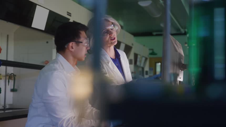 Man and woman wear white lab coats and safety goggles while analyzing data in a laboratory setting