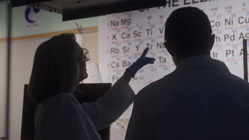The video shows two scientists a man and a woman in lab coats discussing information in a darkened labthe woman is partially lit and is pointing with a bluegloved hand to a large backlit periodic table of the elements on the wall be