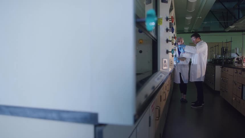 A male and female lab scientist both wearing lab coats gloves and goggles work together in the laboratory focused on their collaborative research
