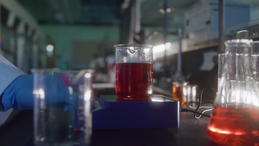 The video shows a beaker of red liquid stirring on a magnetic stirrer in a laboratory settinga person wearing blue gloves is visible in the lowerleft background adjusting another piece of glassware on the lab bench