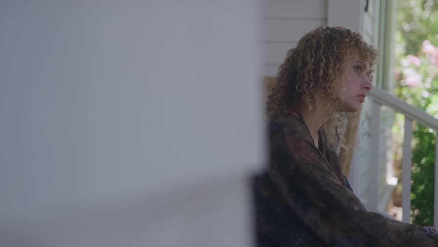 The short video shows a closeup of a young woman with curly lightcolored hair sitting on a porch she is looking offcamera to the right with a serious or thoughtful expressionthe scene is filmed from a slightly obscured angle with a large blurry