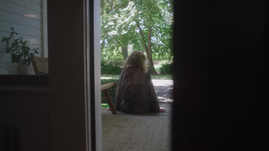 Woman wearing a jacket sitting down on the floor of a porch outdoors during day time