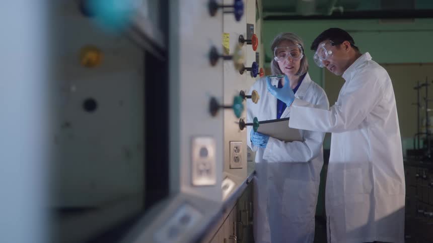 A woman holds a vial as she and a man both wearing white lab coats safety goggles and blue gloves discuss a procedure next to laboratory equipment