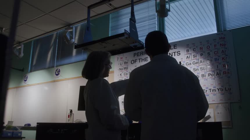The video shows the silhouettes of two people a man and a woman in lab coats facing a brightly lit periodic table of the elements on the back wall of a labthe woman is pointing towards the chart while speaking to her colleague likely