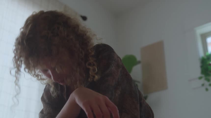 Curlyhaired woman looks thoughtfully at something in a room lit by natural light
