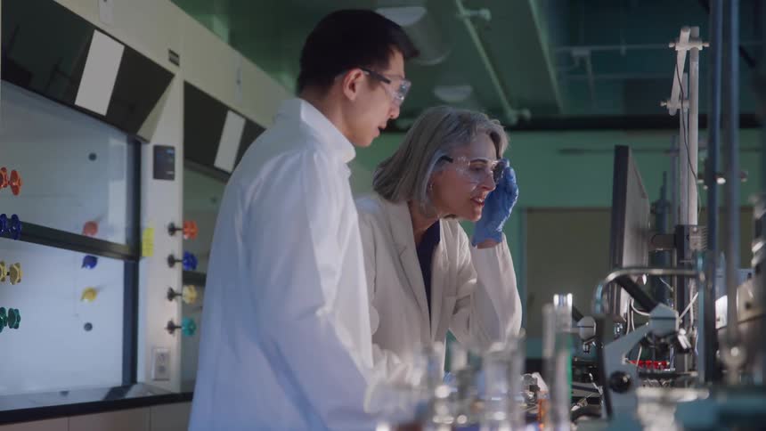 Man and woman wear white lab coats and safety goggles while observing data in a laboratory with a fume hood in the background
