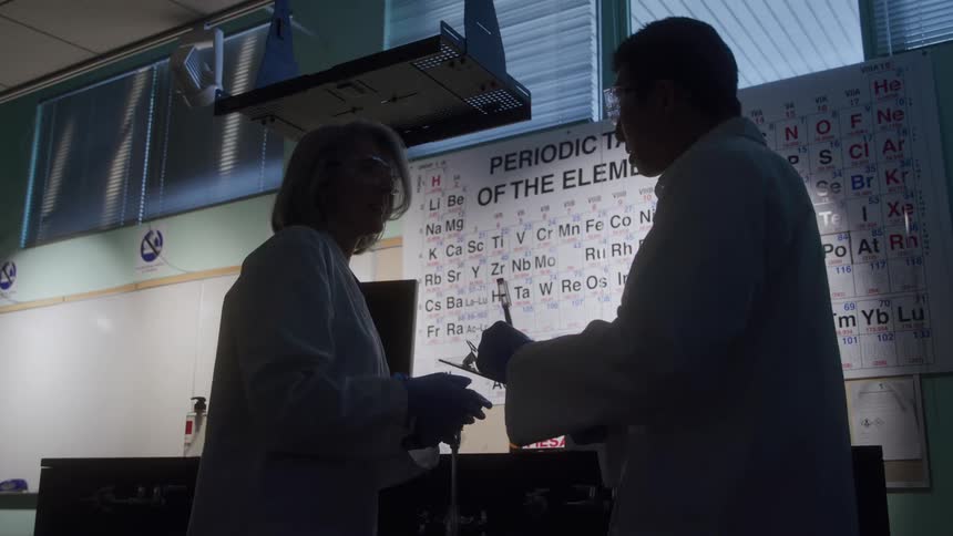 A man and a woman wearing lab coats and safety glasses are silhouetted against a white board while conducting an experiment with test tubes in front of a periodic table chart in a dark science laboratory