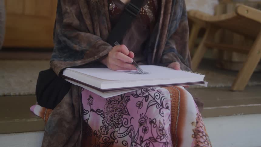 A close view of a woman wearing a decorative brown and pink shawl is seen as she sketches on a white pad of paper with a pencil in an indoor setting