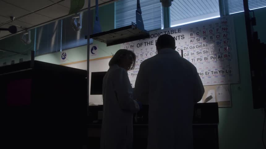 Two scientists in lab coats work in a bluelit laboratory in front of the periodic table
