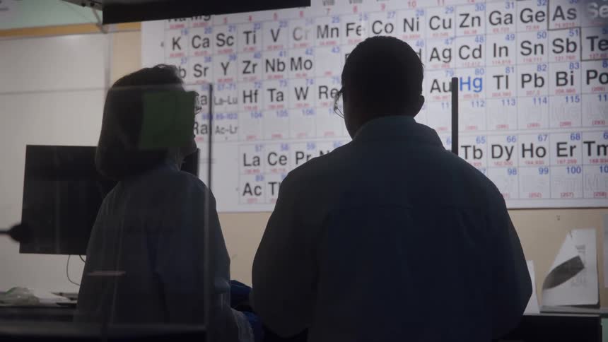 A man and a woman in white coats stand silhouetted with their backs to the camera discussing a project in front of a large brightly lit periodic table