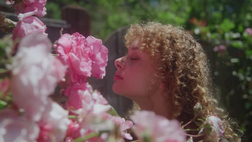 A woman with long curly blonde hair slowly leans in and smells a large cluster of vibrant pink roses in a sunny outdoor garden setting