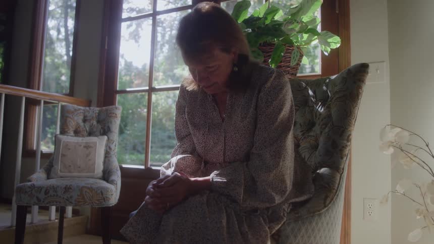 The video shows a woman with reddishbrown hair sitting in a patterned armchair next to a large bright windowshe is looking down at her hands which are clasped in her lap with a pensive or troubled expression