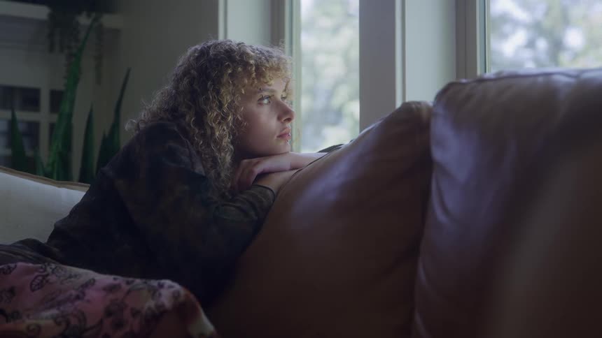 A woman with curly blonde hair leans back on a large brown leather couch and looks out of a bright window at a green outdoor scene