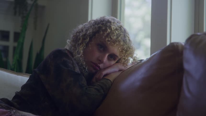A young woman with curly blonde hair sits on a brown couch by the window gazing out with a hint of sadness in the midday light