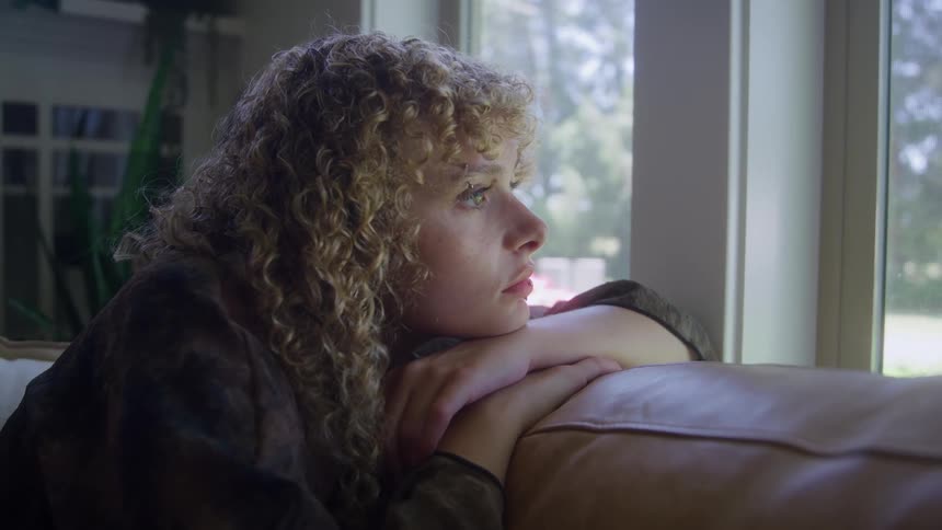 Close up portrait of a young woman looking through a window contemplatively while sitting in a living room during daytime