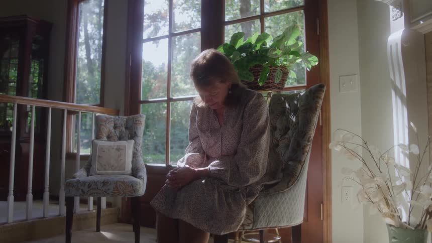 An older woman in a longsleeved floral dress sits alone in a velvet wingback chair with her head down in front of sunlit french doors looking out to a bright green forest