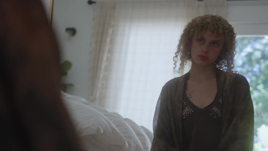The woman sits on the bed by the window