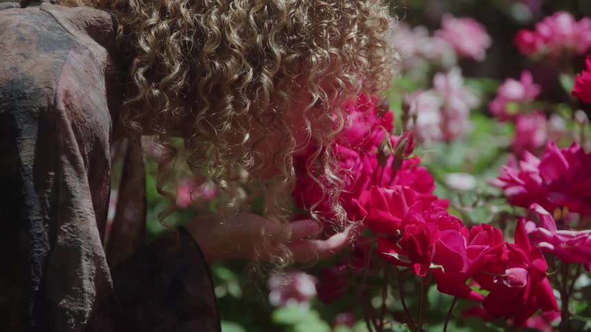 A woman with long curly blonde hair and a dark shirt bends down to gently smell a cluster of vibrant red roses in a sunlit garden full of pink flowers