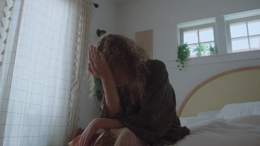 Person sitting on the edge of the bed covering their face with one hand in a gesture of sadness