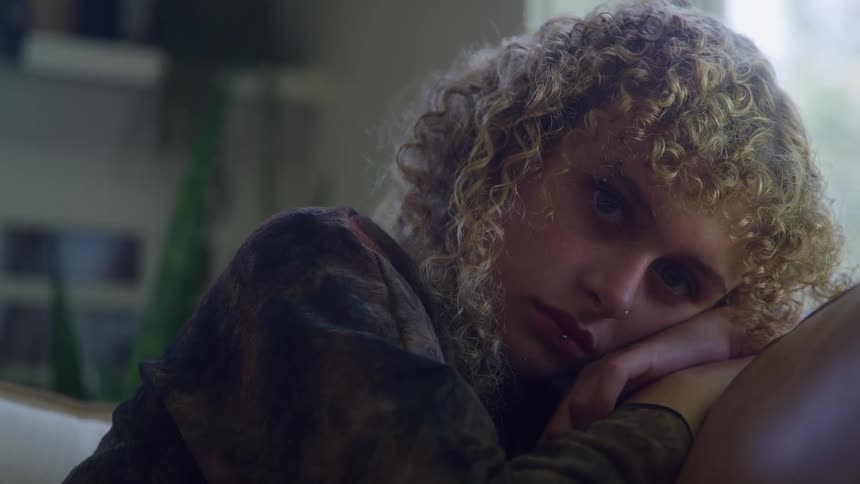 The video is a closeup of a young woman with very curly lightcolored hair resting her chin on her handshe appears to be sitting near a window with her eyes moving offcamera conveying a pensive or sad mood
