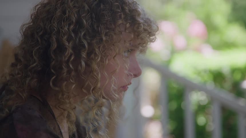 A close view of a woman with long curly blonde hair shows her looking down with a somber expression as she sits outside on a porch railing with a bright green and pink floral background