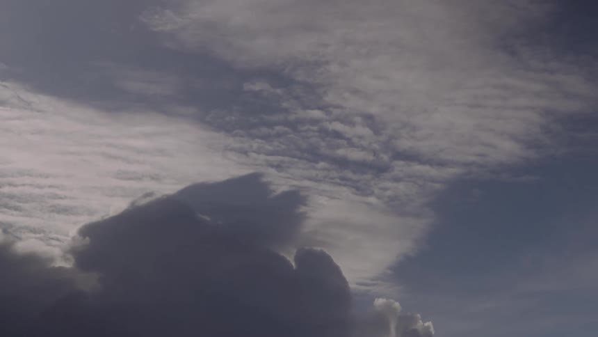 A timelapse view shows a large dark cumulus cloud swelling into a taller formation against a blue sky with soft white cirrus clouds moving across the background