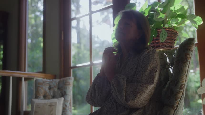 Woman sitting by the window with her hands clasped appearing calm and contemplative
