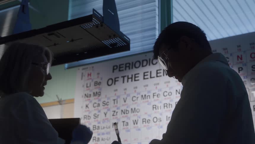 A lowangle silhouetted view shows a woman and a man in lab coats and safety glasses discussing information on a brightly lit periodic table with the man holding a pen